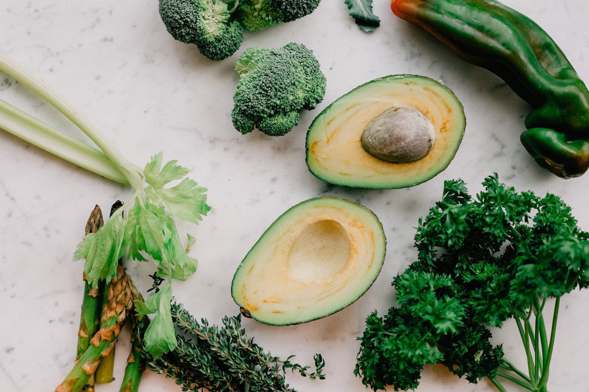 Green vegetables arranged on a white surface: broccoli, avocado halves, asparagus, herbs, and pepper.
