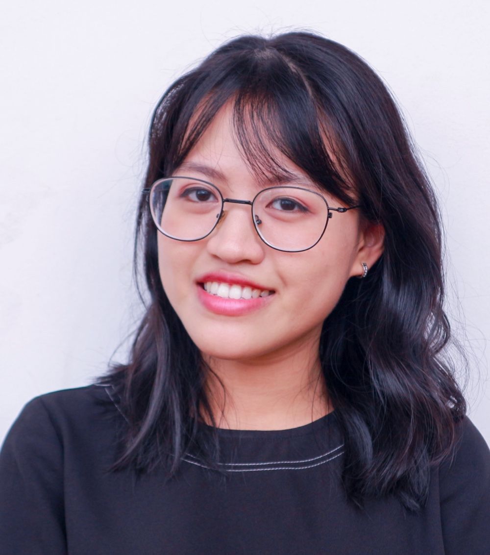 Woman with glasses smiling, looking at the camera. Dark hair, black shirt, white background.