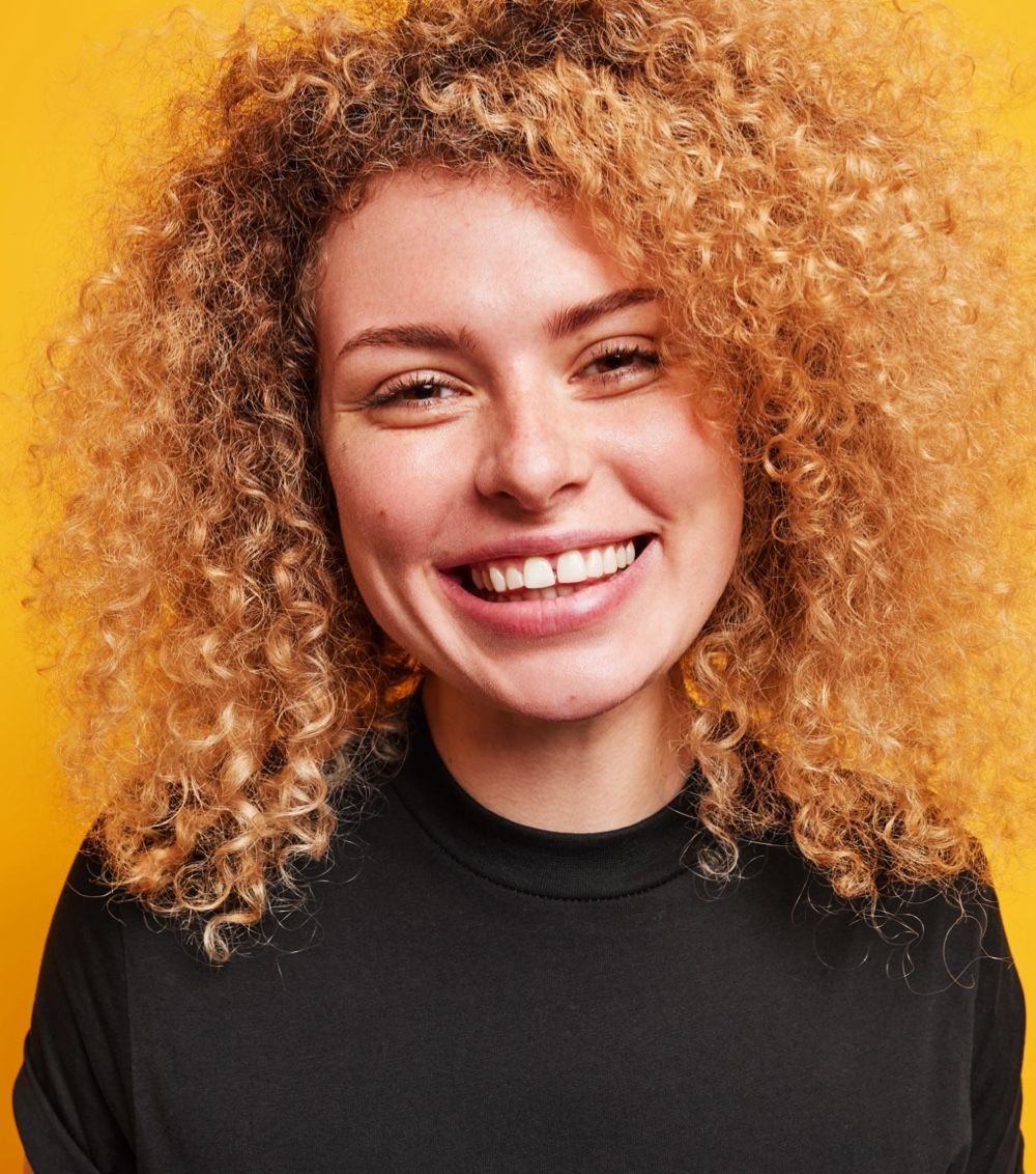 Woman with curly blonde hair smiles at the camera, wearing a black top against a yellow background.