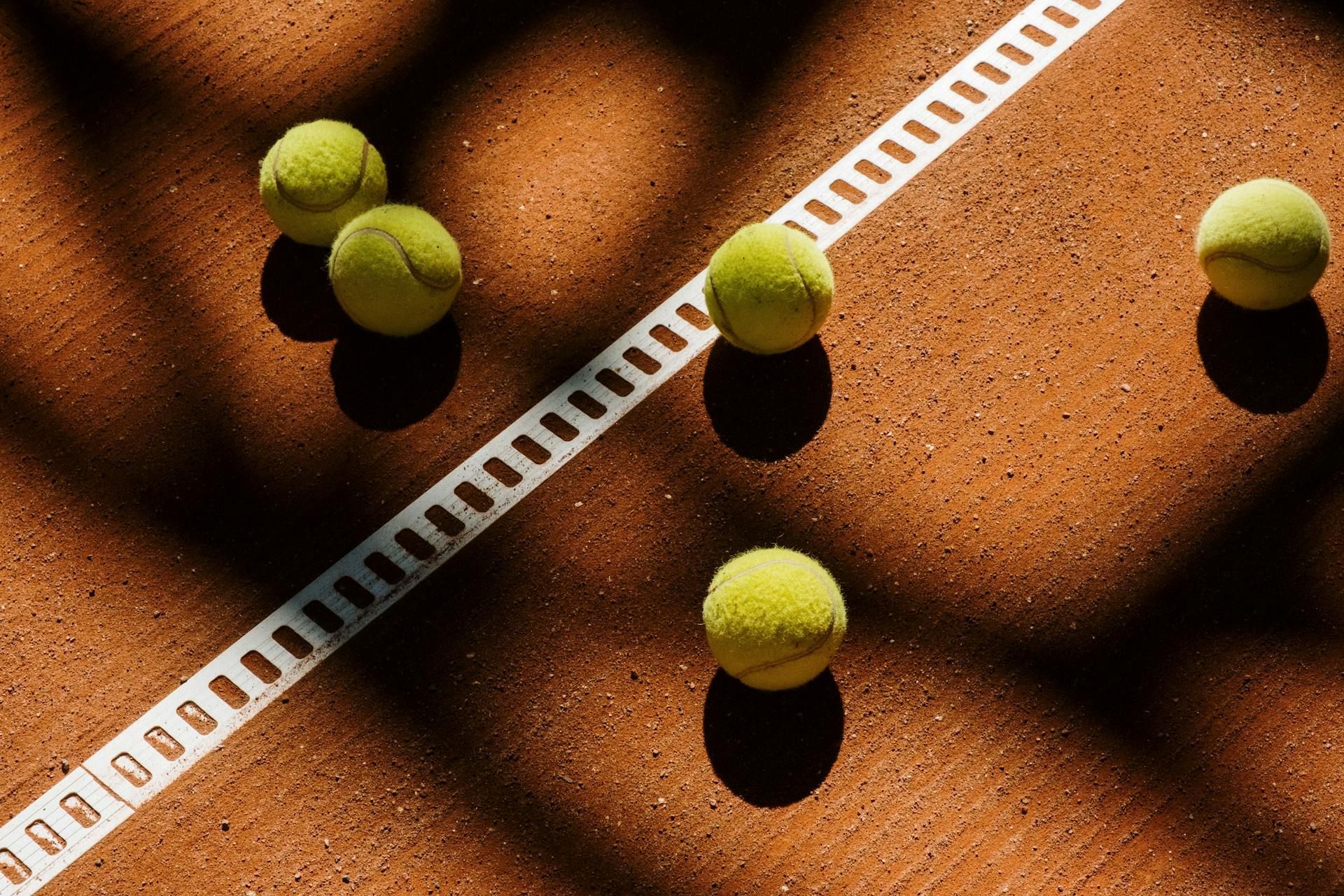 Five yellow tennis balls on a clay court beside a white sideline shadow pattern