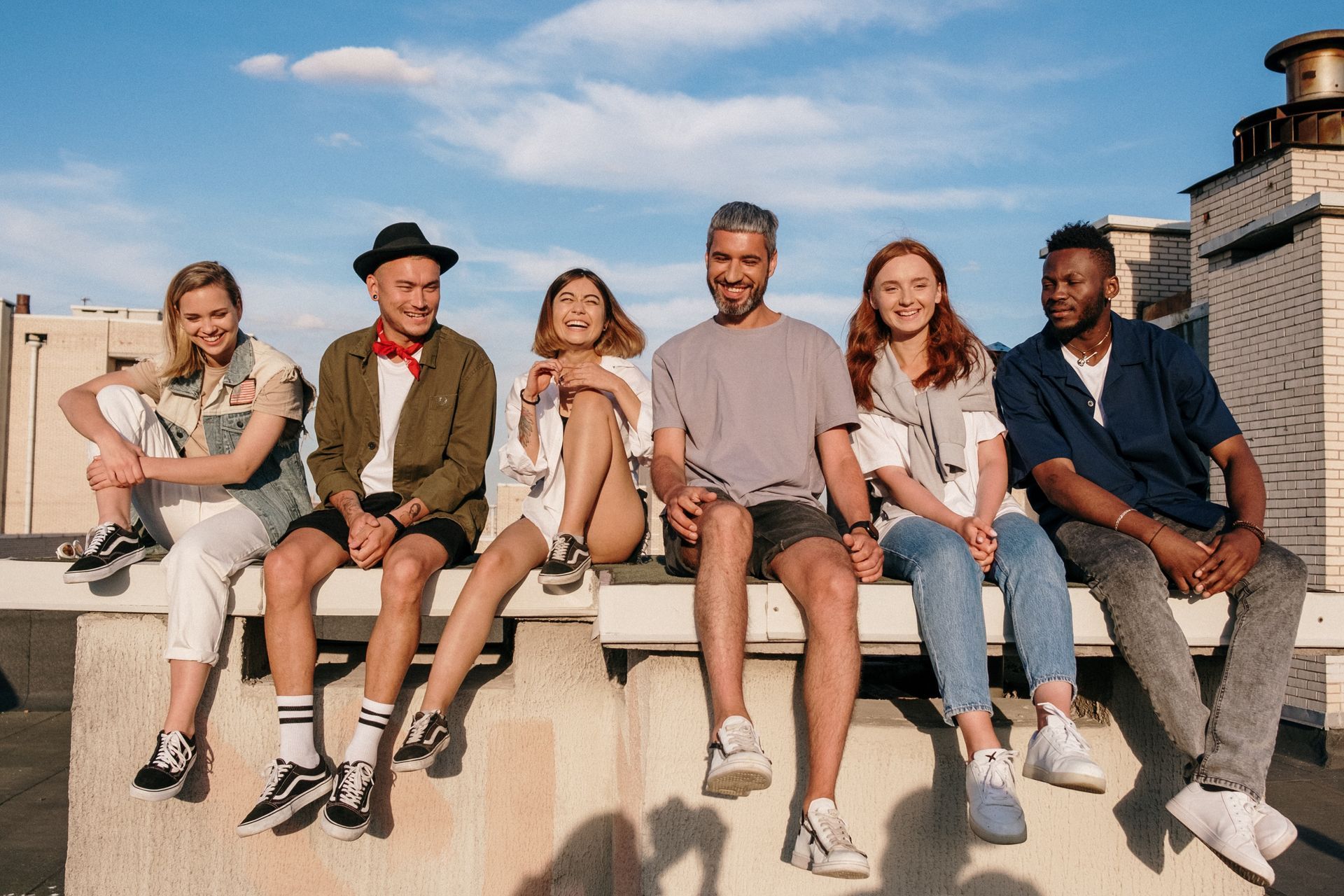 Six friends sit together in a line on a rooftop ledge against a clear blue sky, smiling and looking toward the camera.