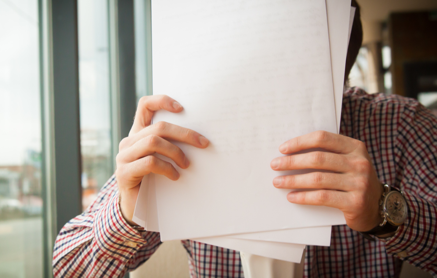 Person holding stack of white papers in front of their face, covering it. Wearing a checkered shirt, watch, near a window.