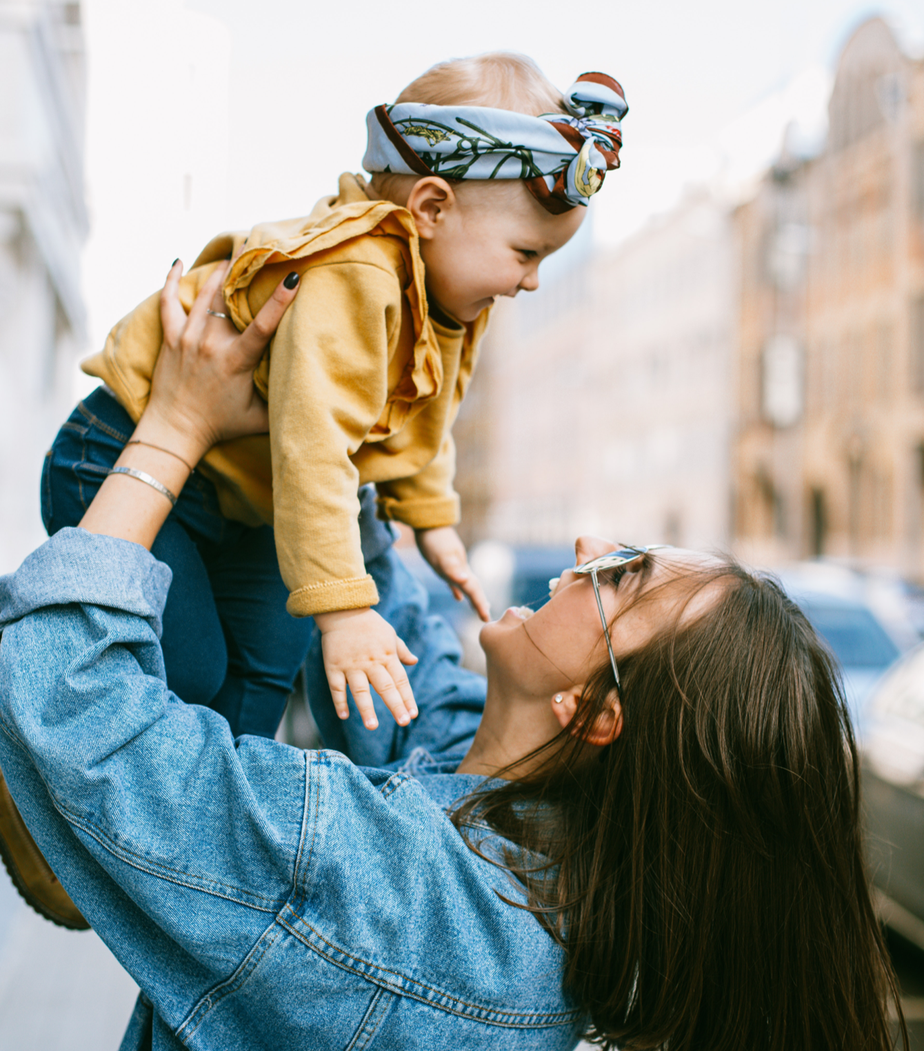 Woman in denim jacket holds a baby up in the air, both smiling, outdoors.