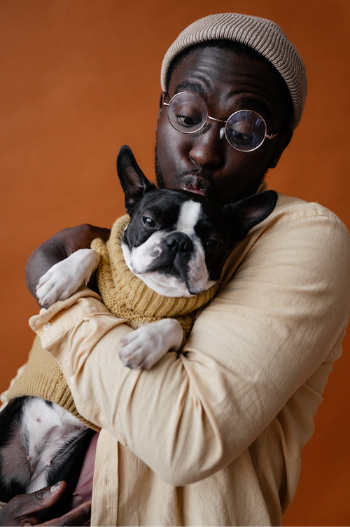 Person in a beige sweater and hat cuddles a black-and-white dog against an orange background