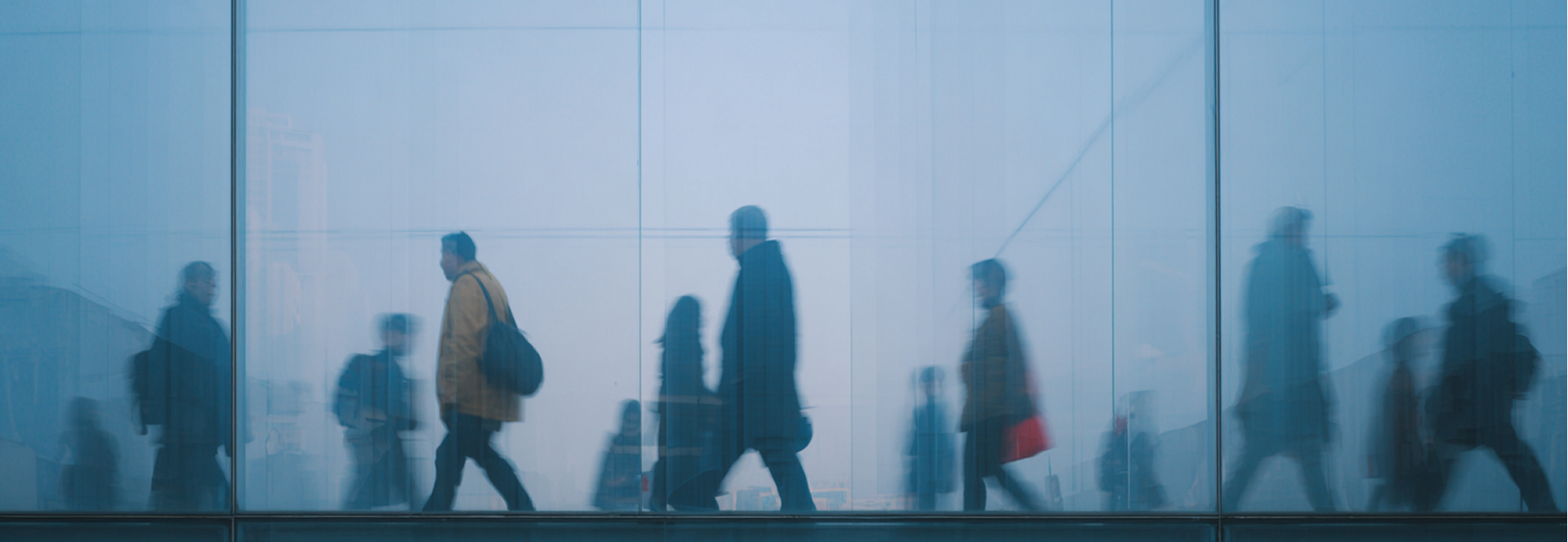Silhouetted figures walking past a glass wall. Overcast day with a blue hue.