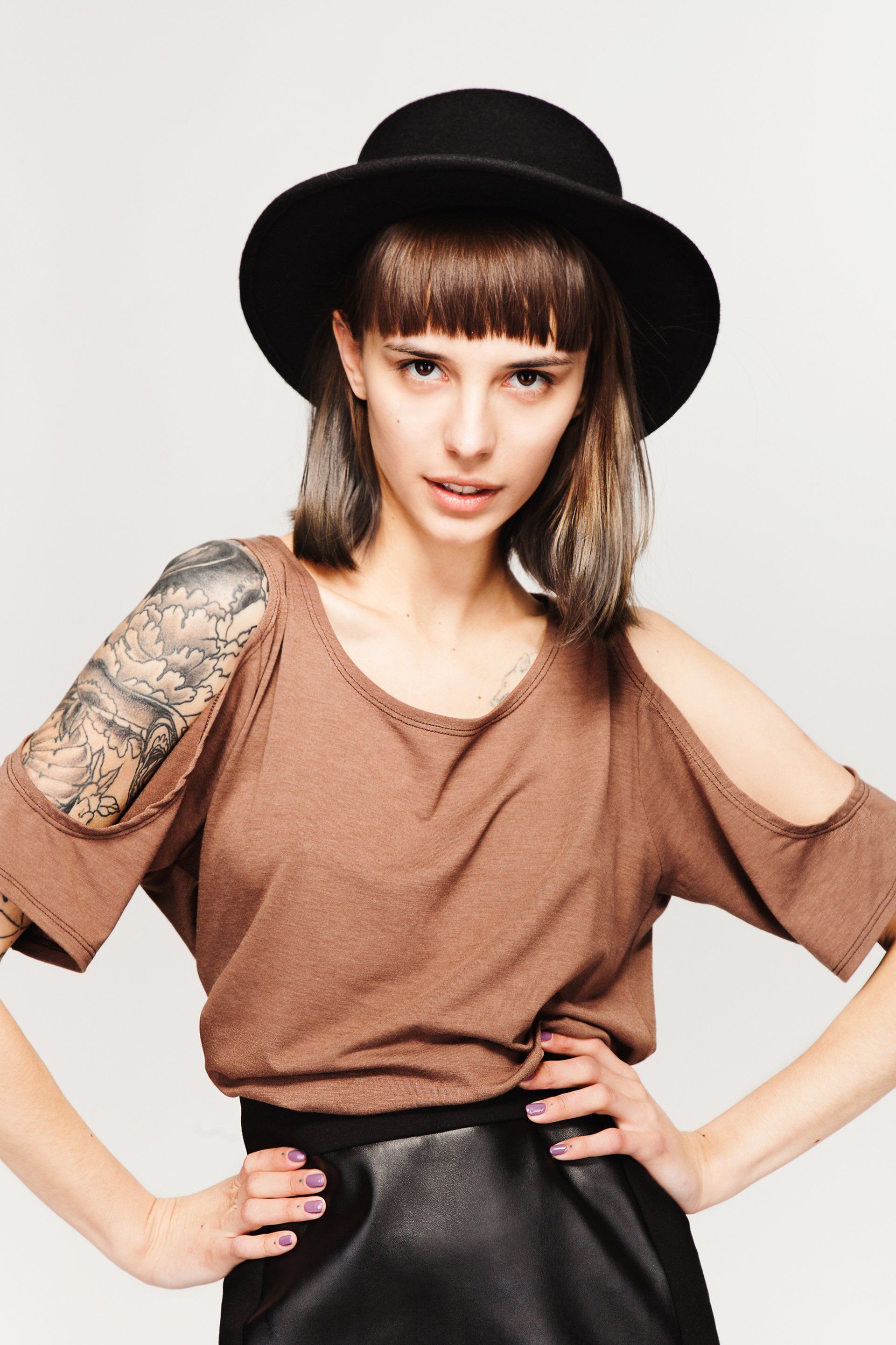 Woman in brown top, black skirt, and hat with arms on hips, posing in studio.