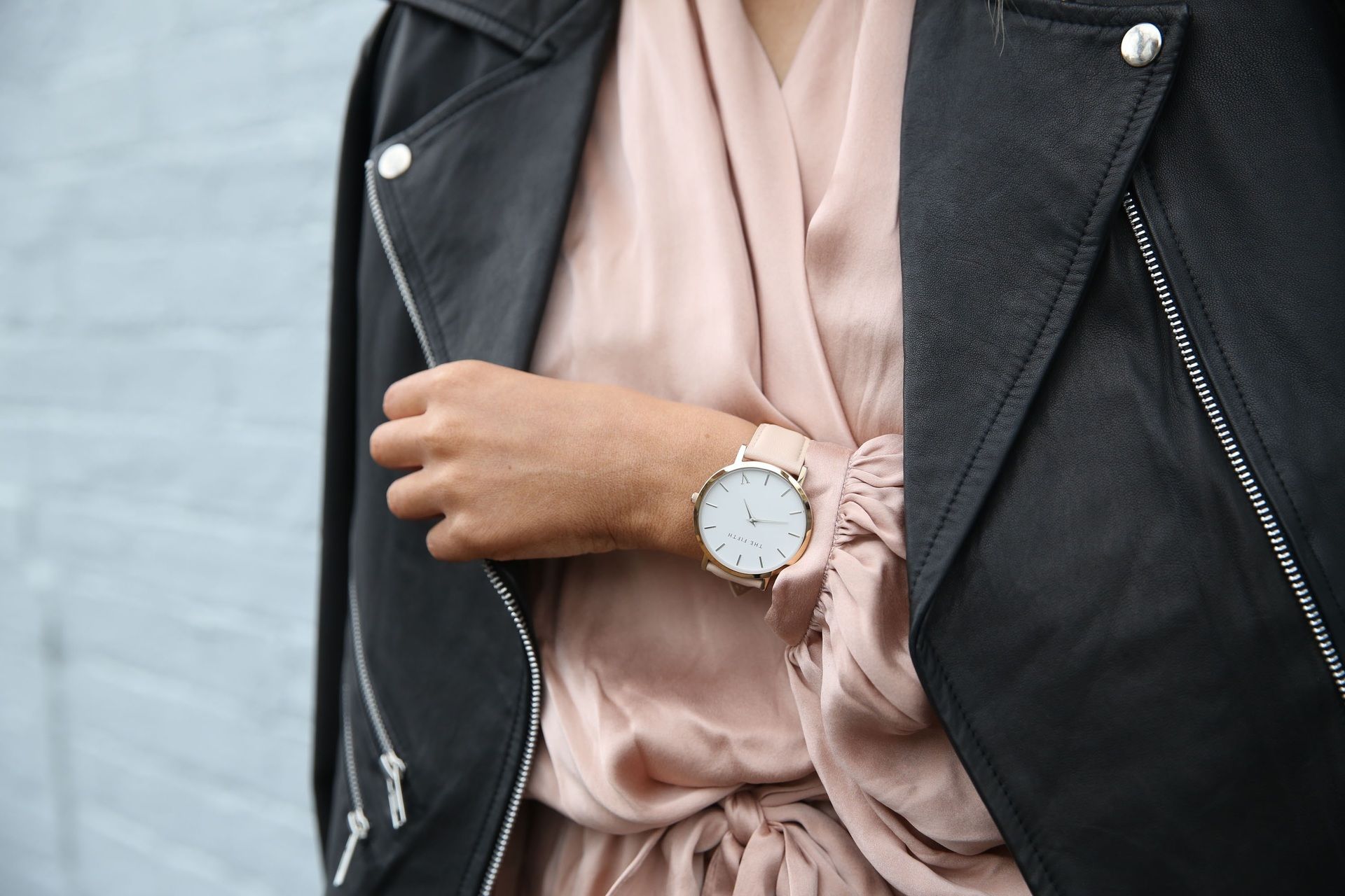 Woman wearing a black leather jacket, pink satin top, and silver watch, standing in front of a gray wall.
