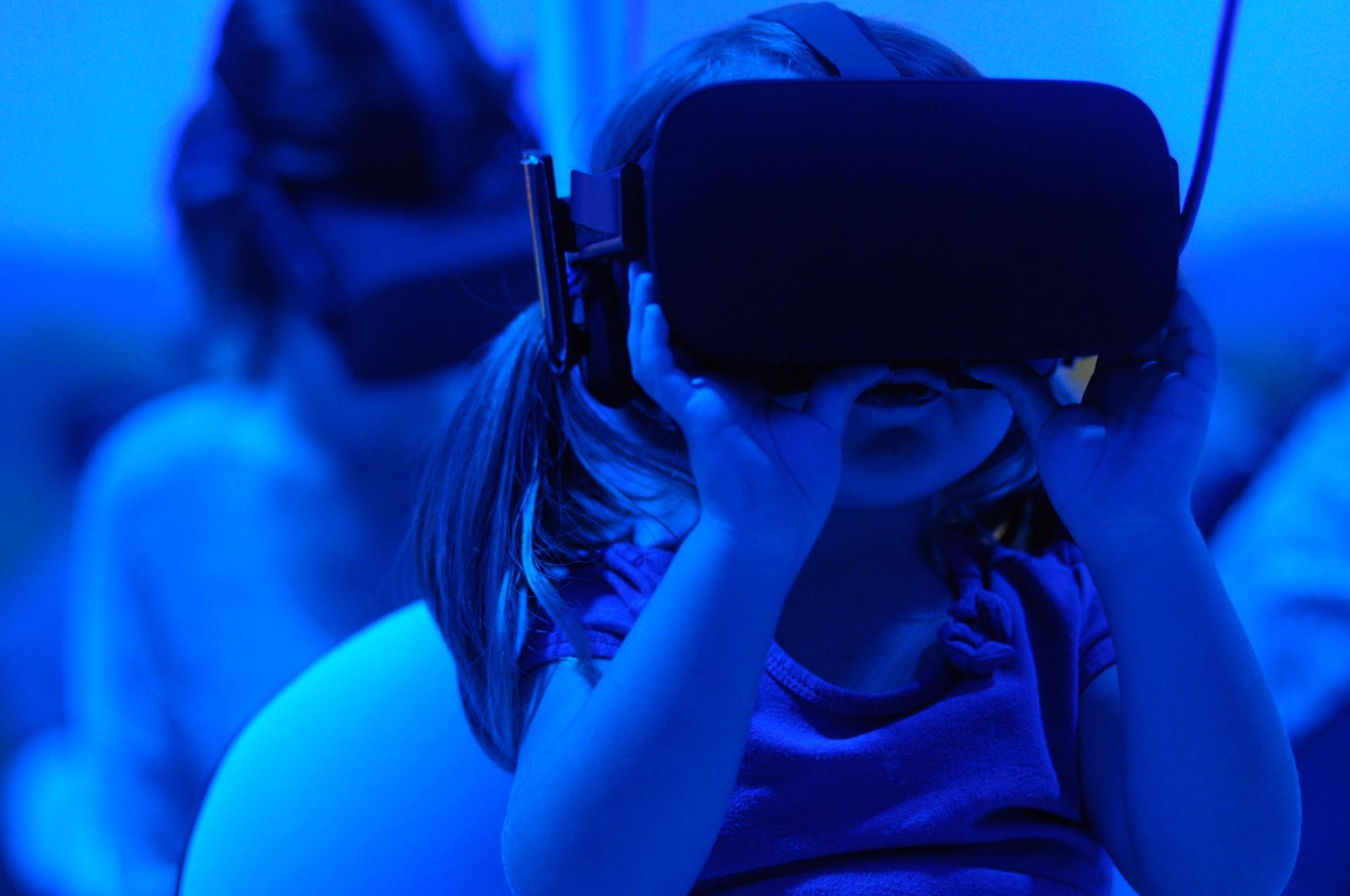 Girl wearing VR headset, looking up, illuminated by blue light. Another person in background.