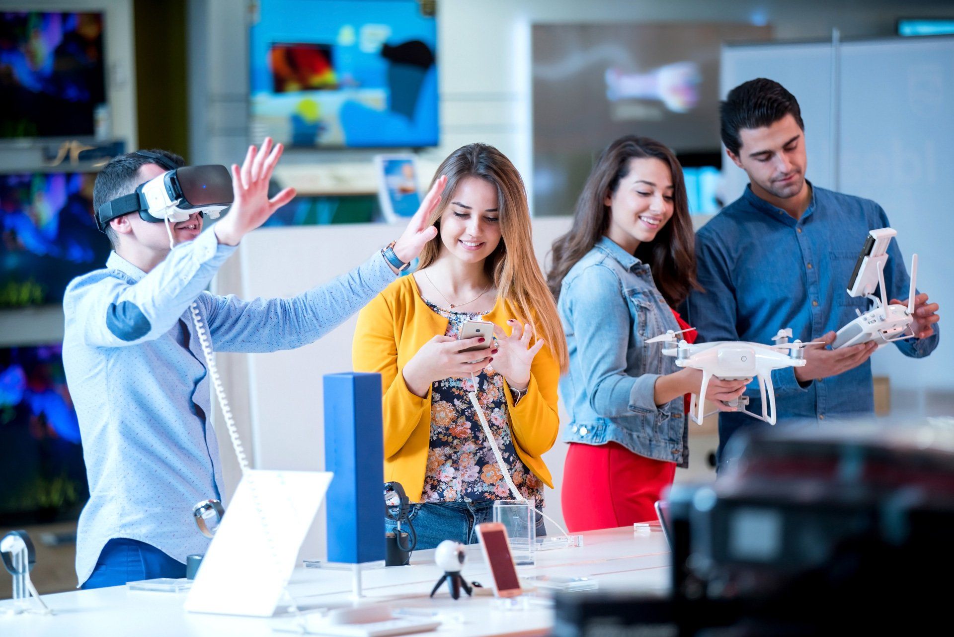 People in an electronics store: one wearing VR headset, others looking at a drone and phones.