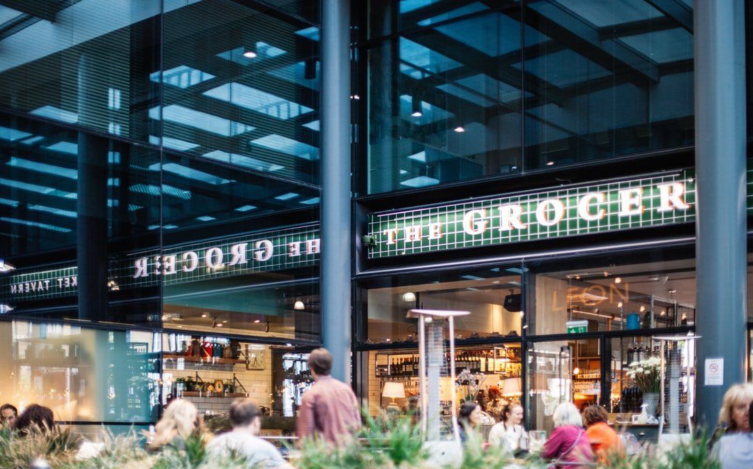The Grocer restaurant with glass facade, featuring a busy outdoor seating area and sign.