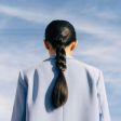 Person seen from behind with long braided hair wearing a light blue shirt against a plain background