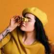 Person in a yellow beret and sweater holds a yellow flower to their face against a yellow background.