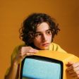 Person with curly hair holding a yellow retro TV monitor against a black background