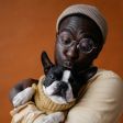 Person in a gray hat and round glasses holding a black-and-white cat against an orange background