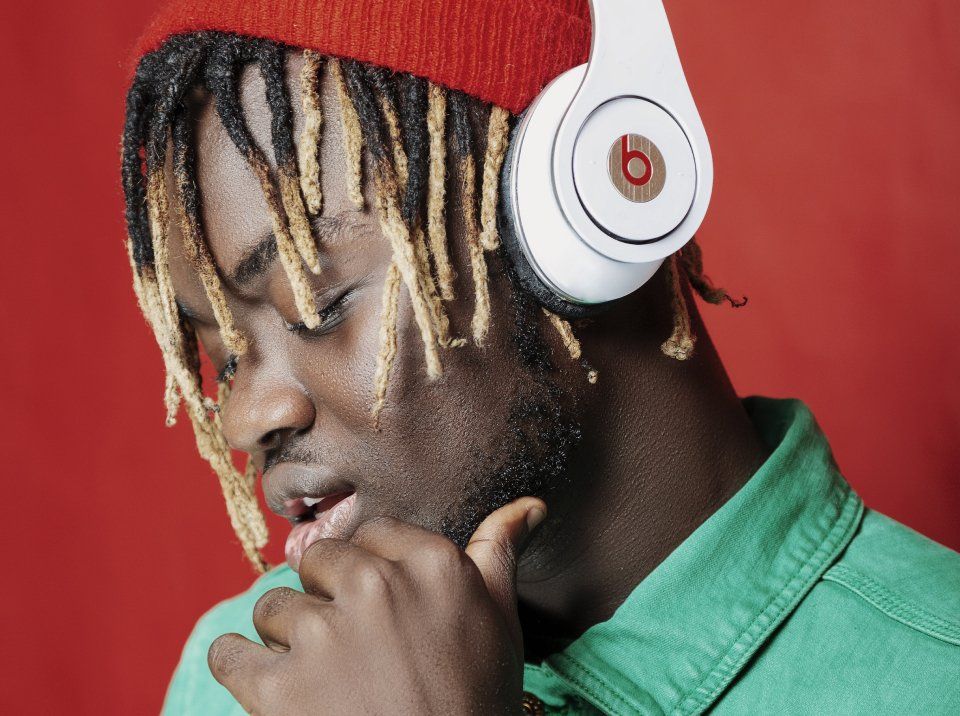 Man wearing white headphones and a red beanie, looking down with a contemplative expression against a red background.