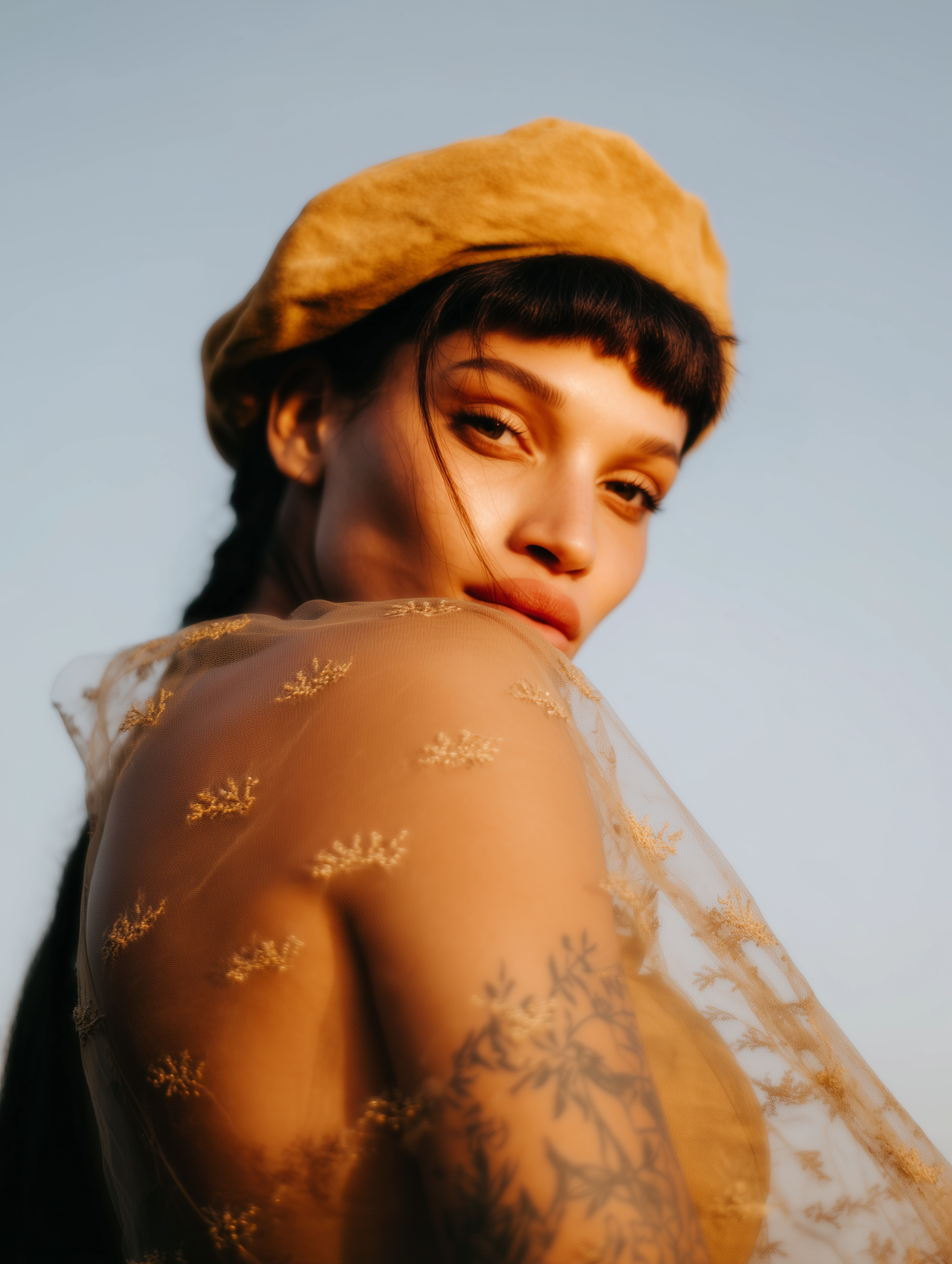 Woman in beret and sheer top, looking over shoulder. Golden sunlight illuminates skin and floral tattoos.