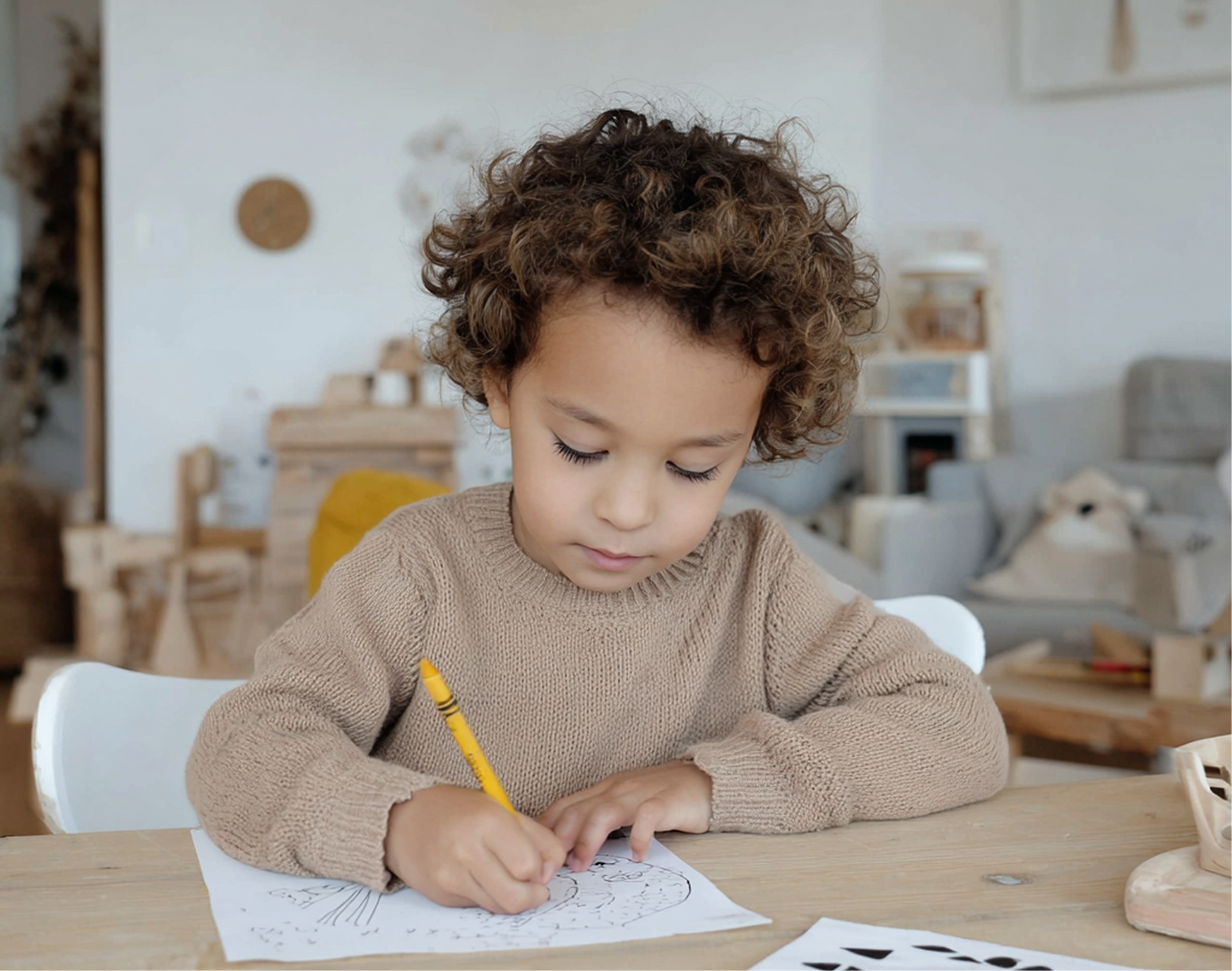 Young child with curly hair drawing with a yellow pencil at a wooden table.