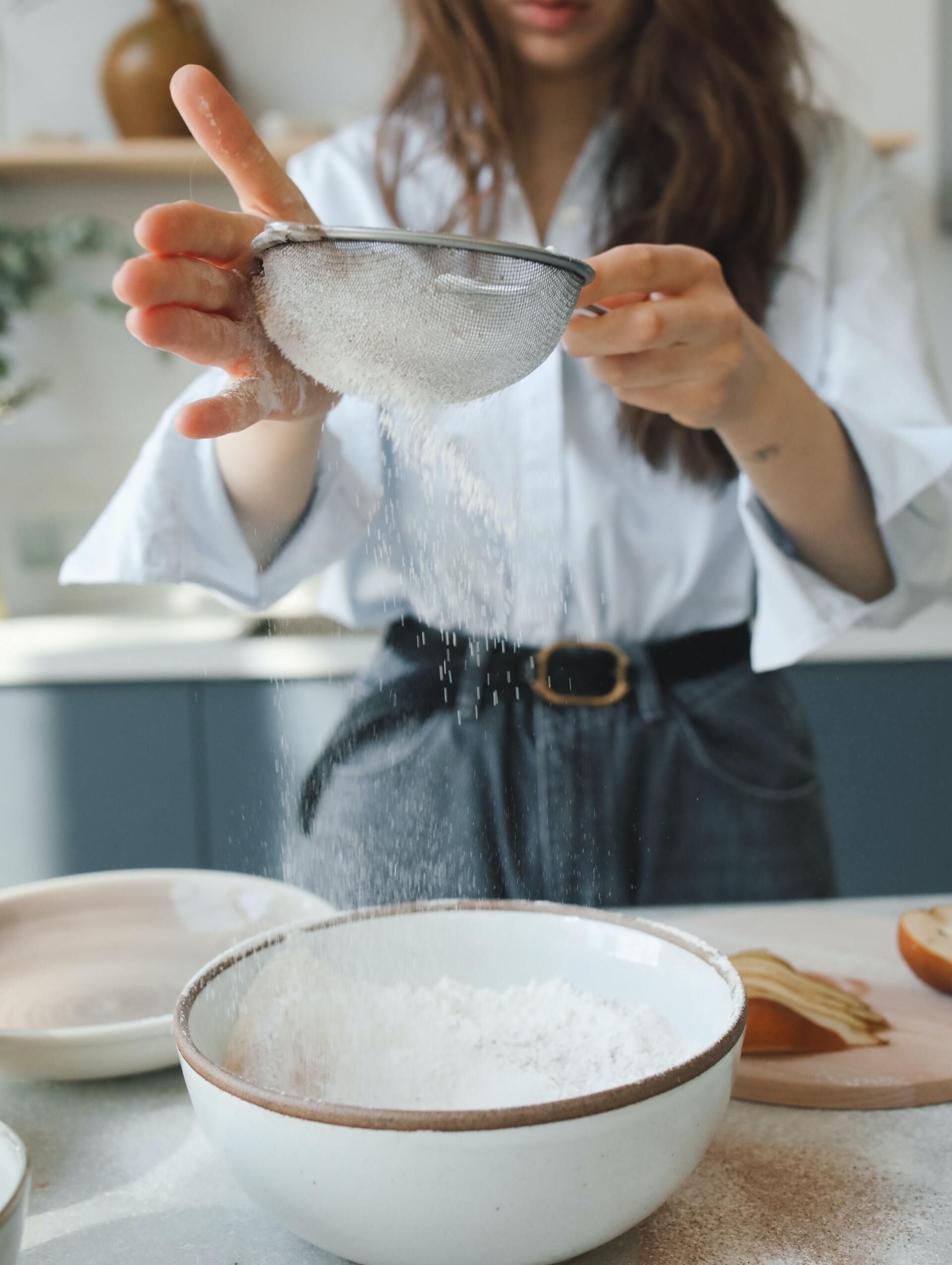 A person in a light shirt sifts white flour into a large ceramic bowl in a kitchen setting.