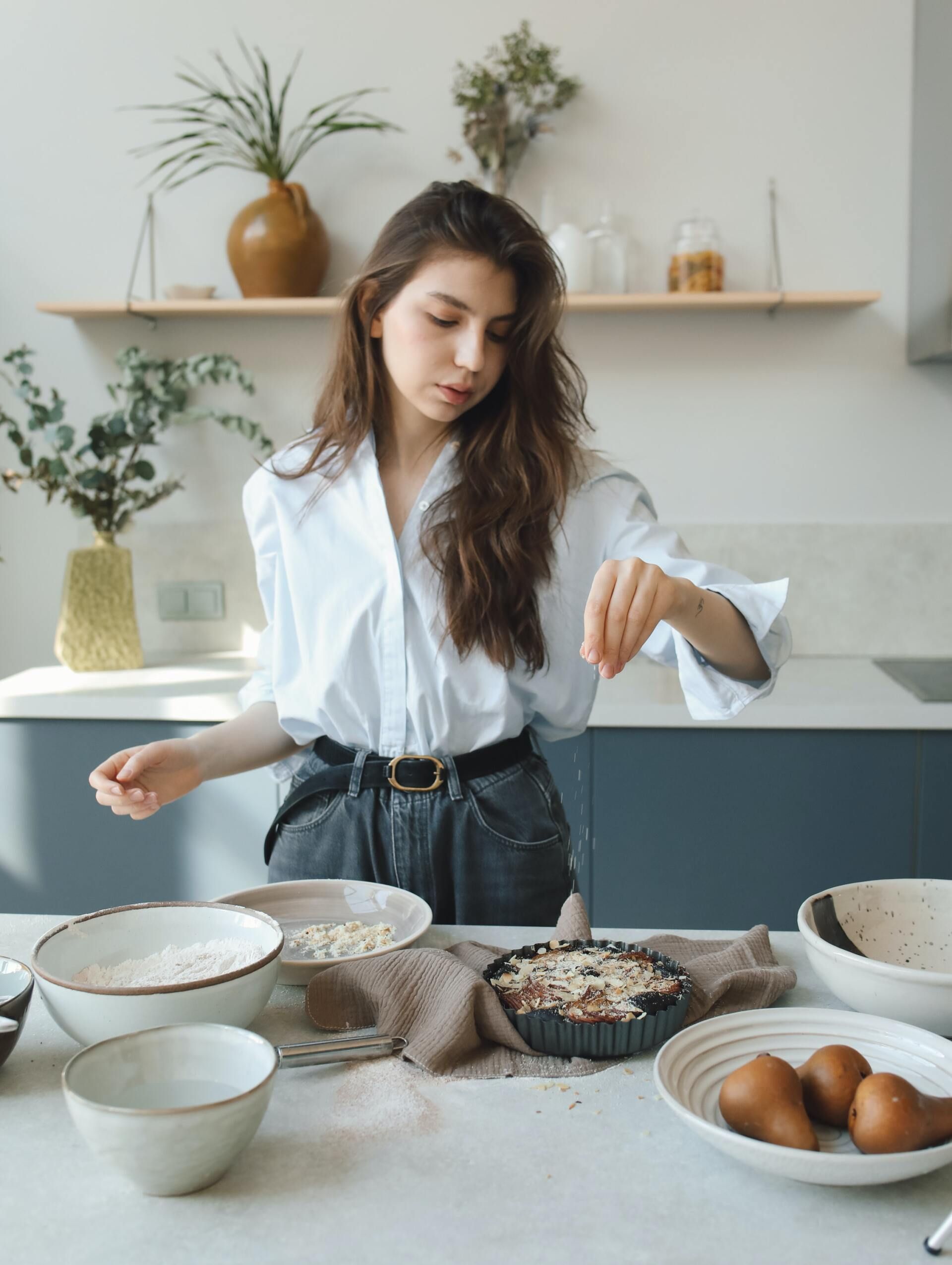 A person in a white shirt prepares food in a kitchen, sprinkling ingredients onto a tart in a baking dish.