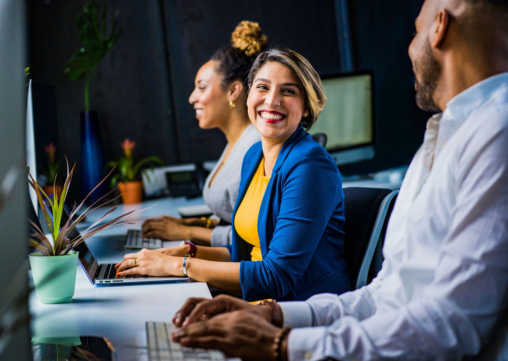 Three coworkers at desks, smiling, working on computers.