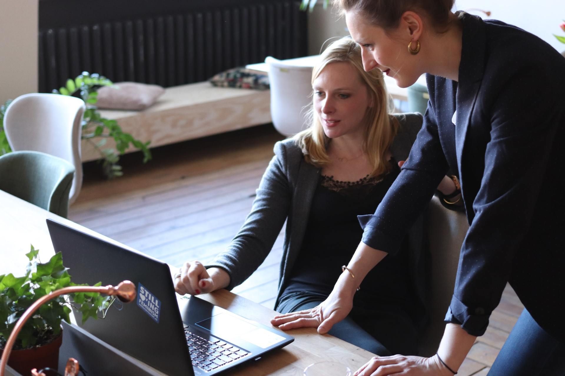 Two women looking at a laptop, one points at the screen. Office setting with plants.