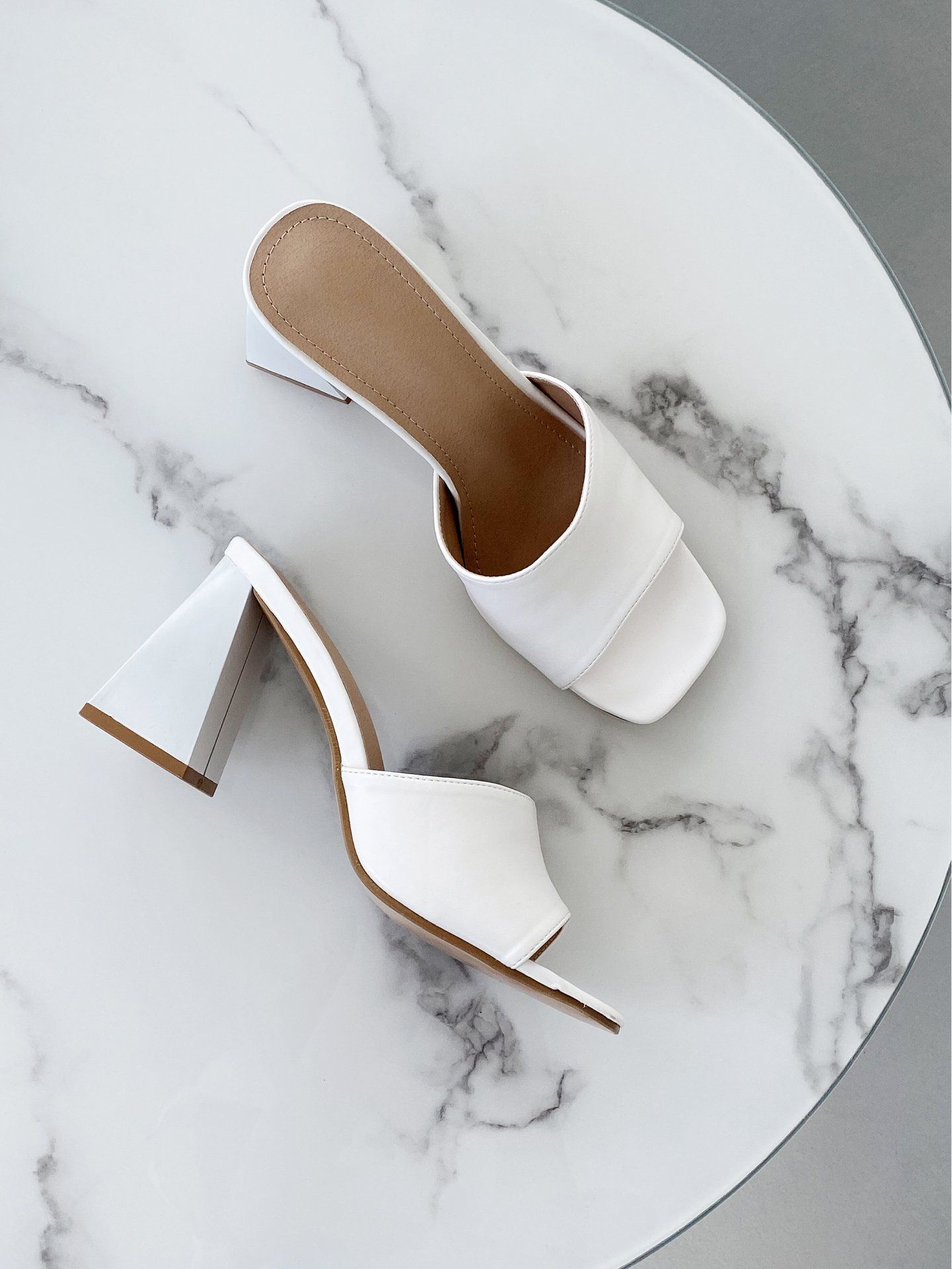 White heeled slide sandals on a marble surface, shown from top and side views.