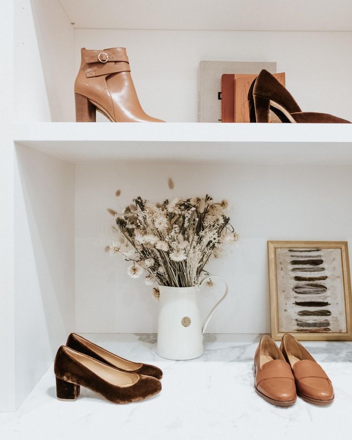 White shelves displaying several pairs of tan and brown heeled shoes, a vase of dried flowers, and framed art