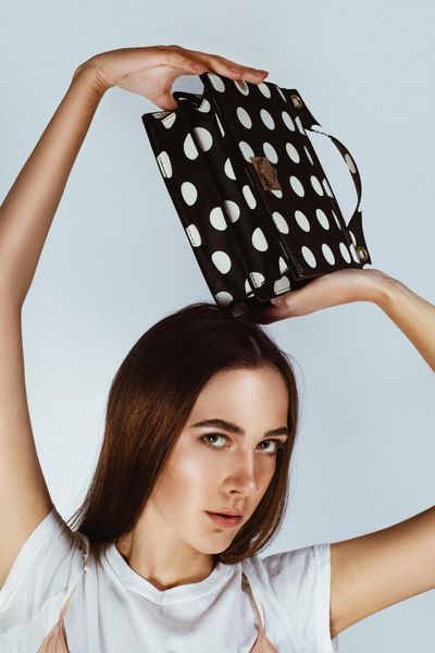 Woman posing with a black-and-white polka dot bag balanced on her head against a plain background