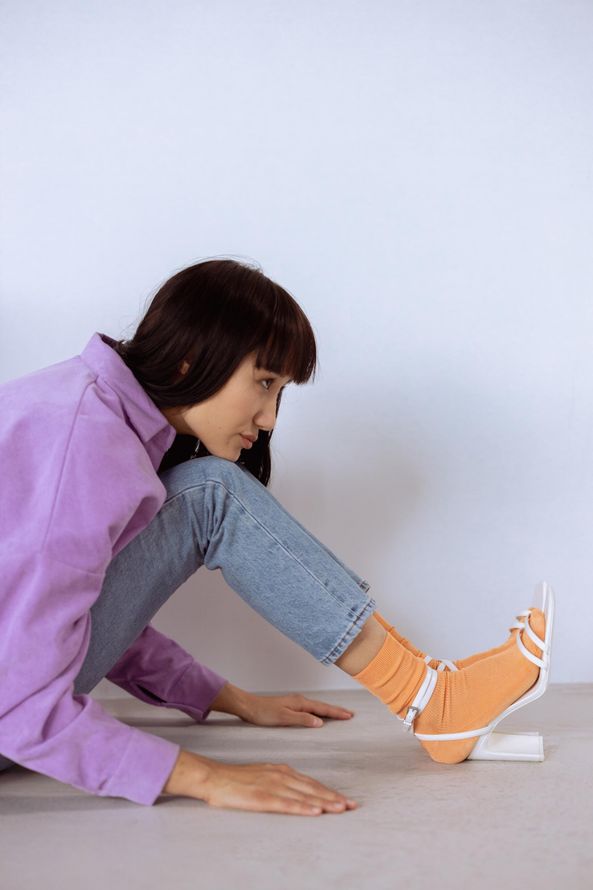 Woman stretching her leg against a wall on a light floor, wearing a purple shirt, jeans, and orange sneakers