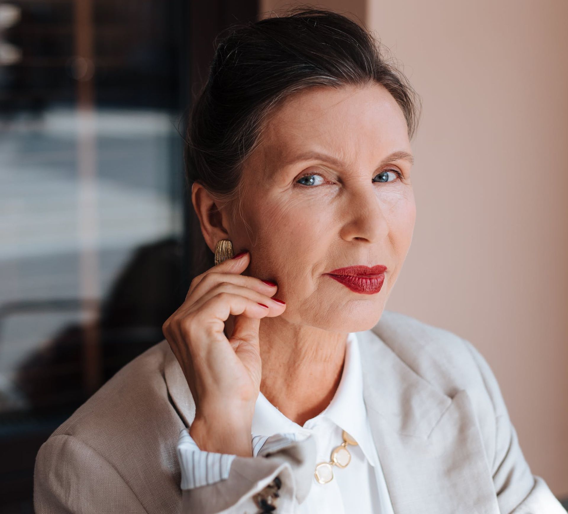 Woman in a white blazer and shirt, red lipstick, looking towards the viewer, holding her cheek.