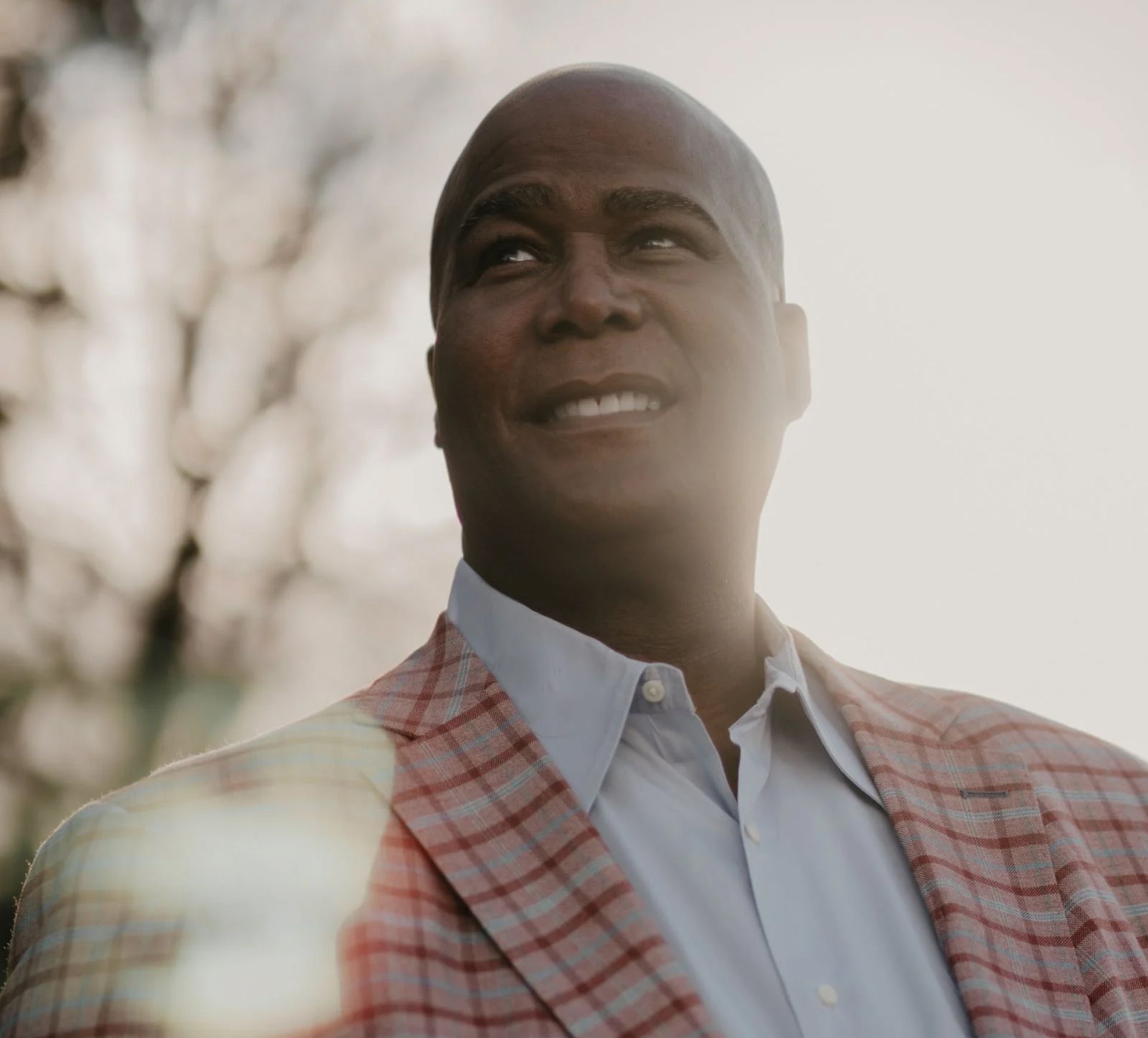 Man in light blue shirt and red plaid blazer looks up and smiles outdoors.