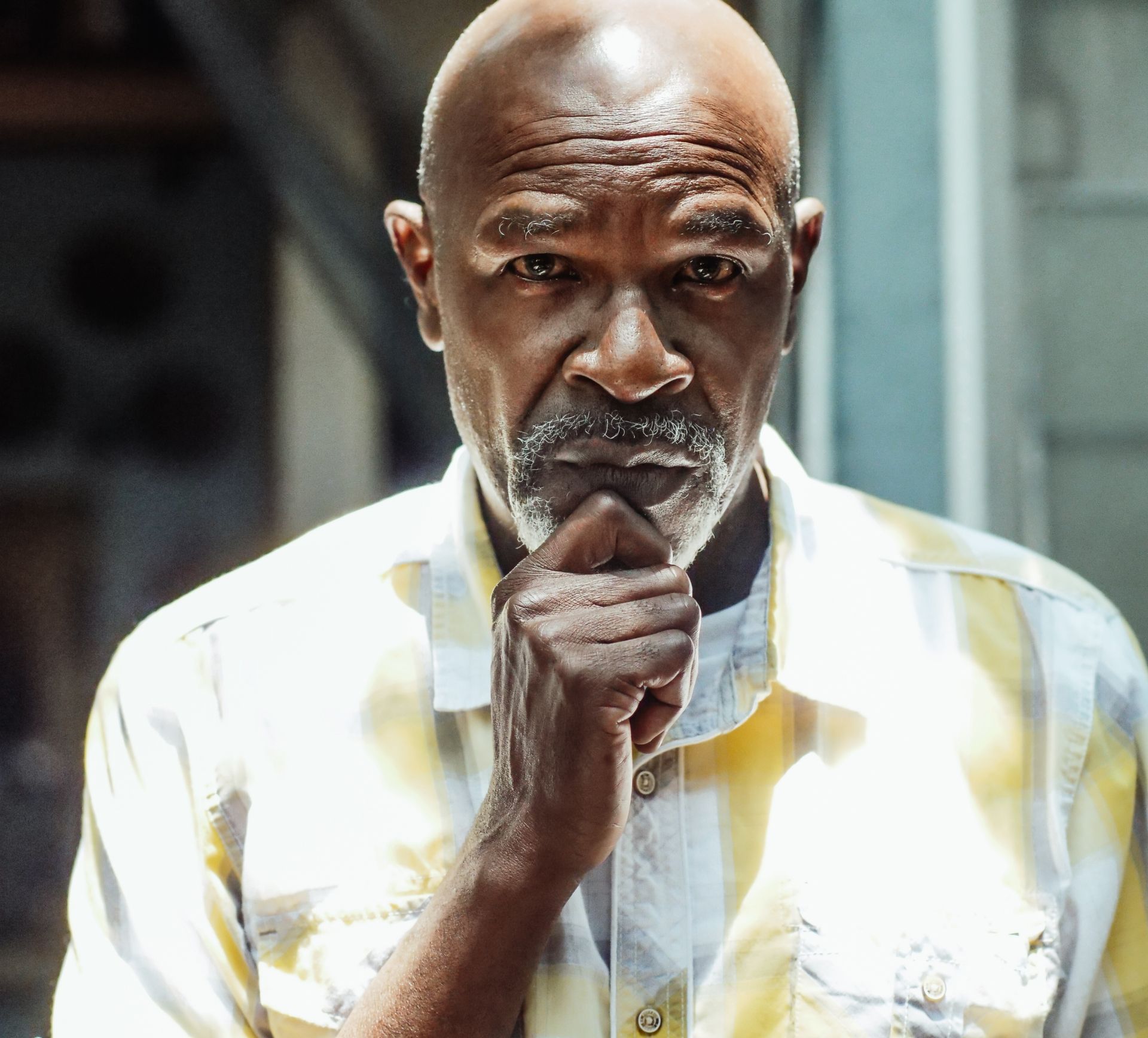 Man with shaved head, hand on chin, thoughtful expression, wearing a yellow-patterned shirt.