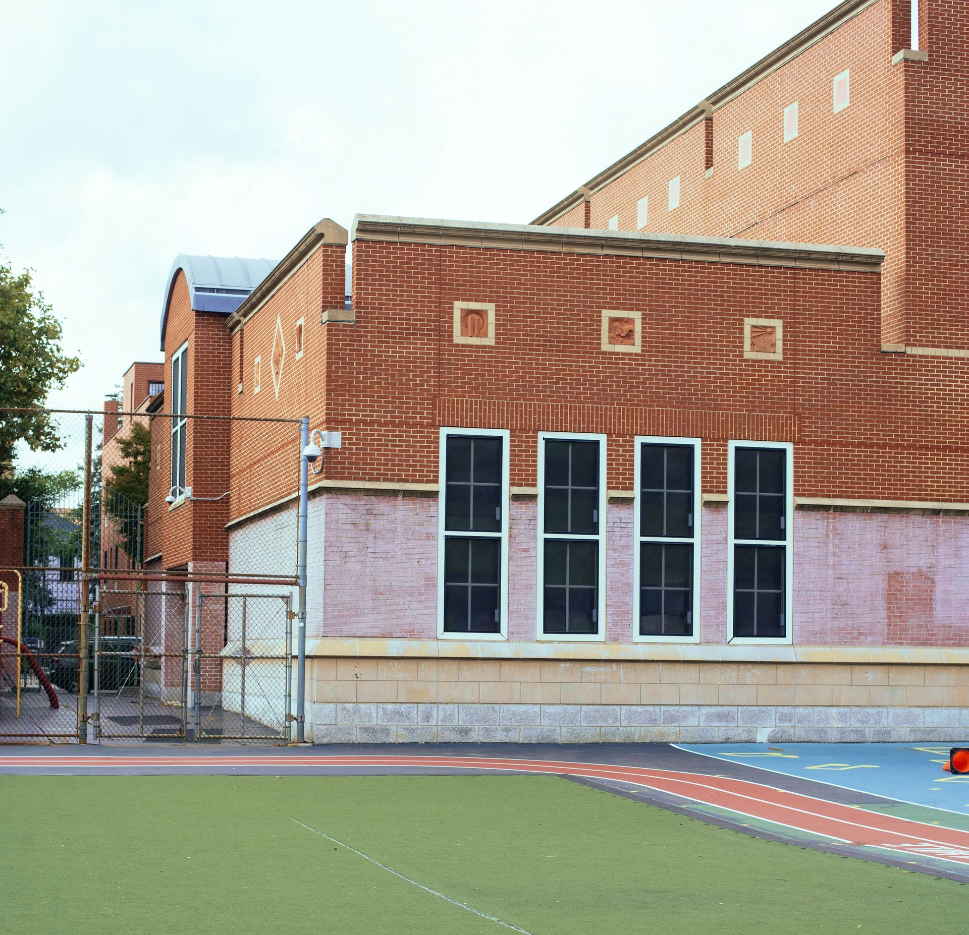 Brick school building with windows, overlooking a green and blue playground, under a cloudy sky.