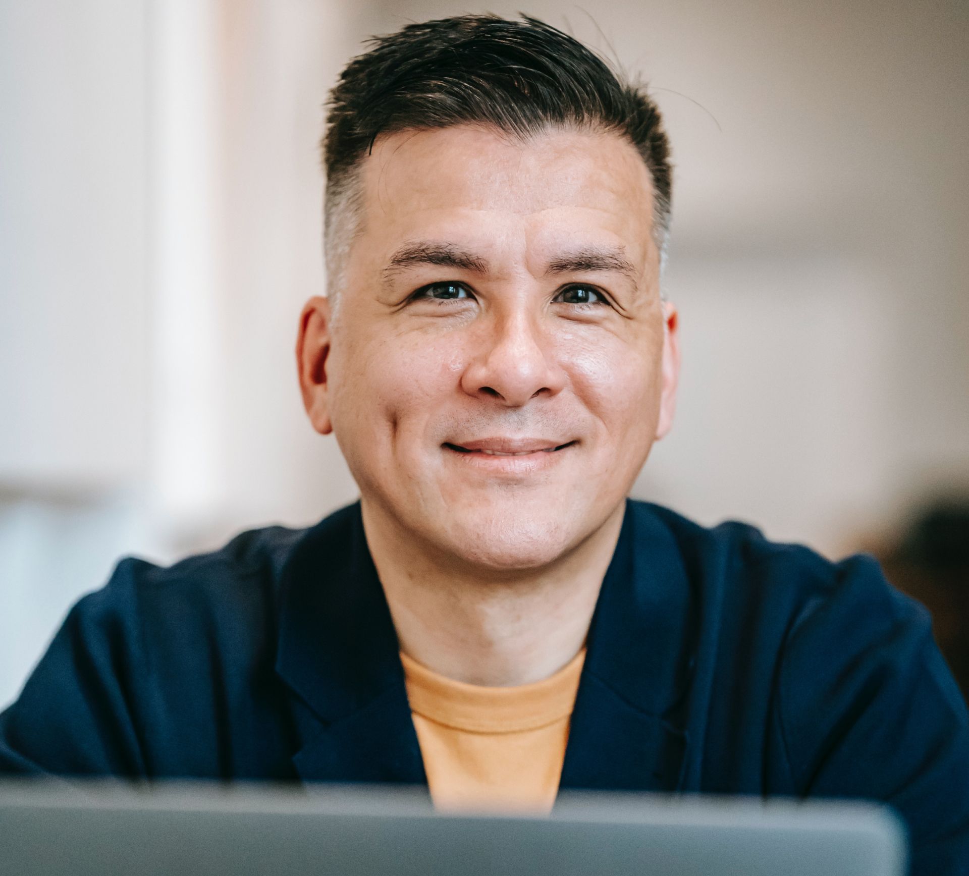 Man with dark hair and a dimple smiles, wearing a navy jacket over an orange shirt, indoors.