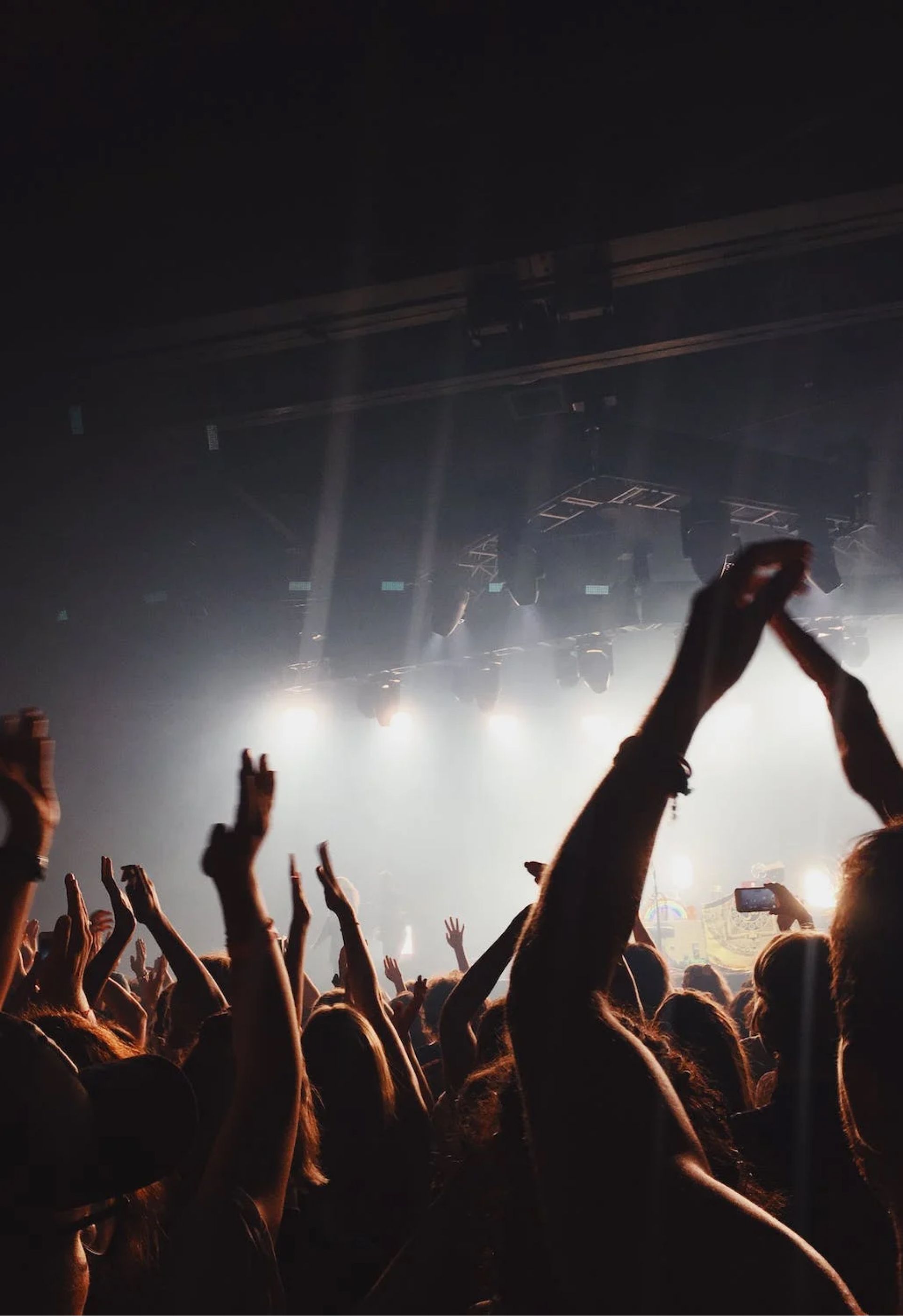A crowd at a concert with their hands raised toward bright stage lights in a dark venue.