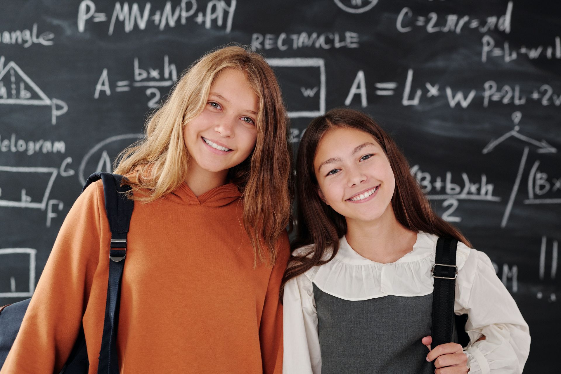 Two smiling students stand in front of a chalkboard covered with handwritten geometric formulas and diagrams.