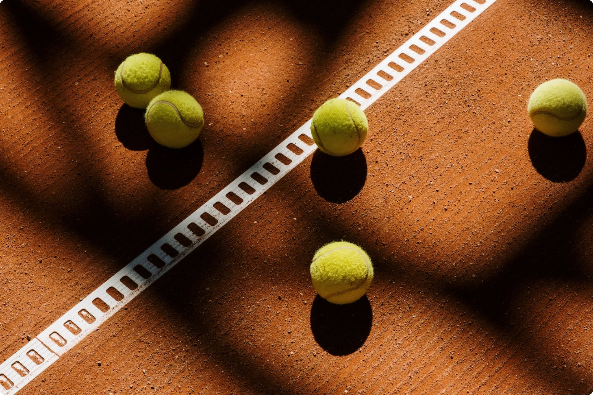 Tennis balls on a clay court near a white line, with shadows cast by the sun.