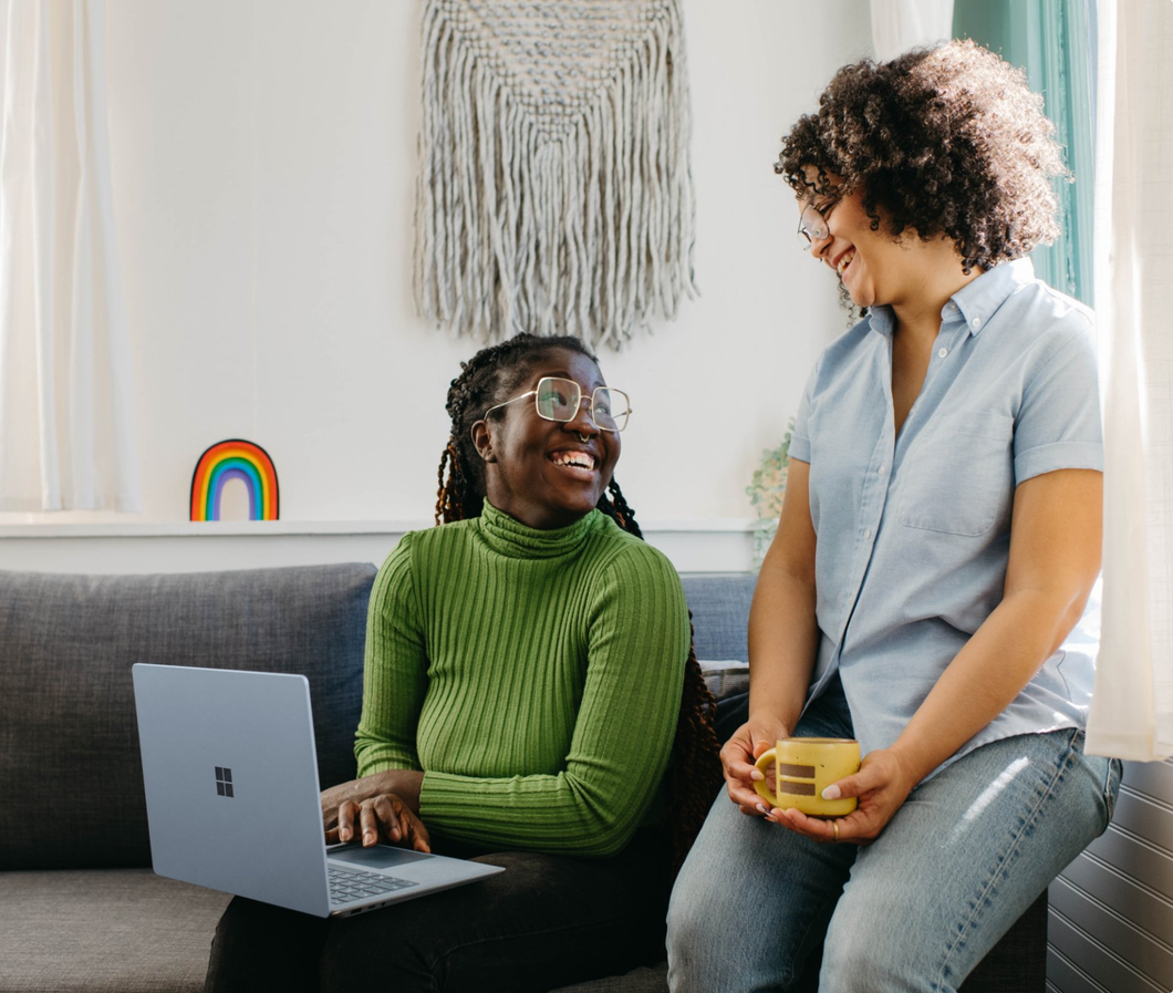 Two women are sitting on a couch with a laptop and a cup of coffee.