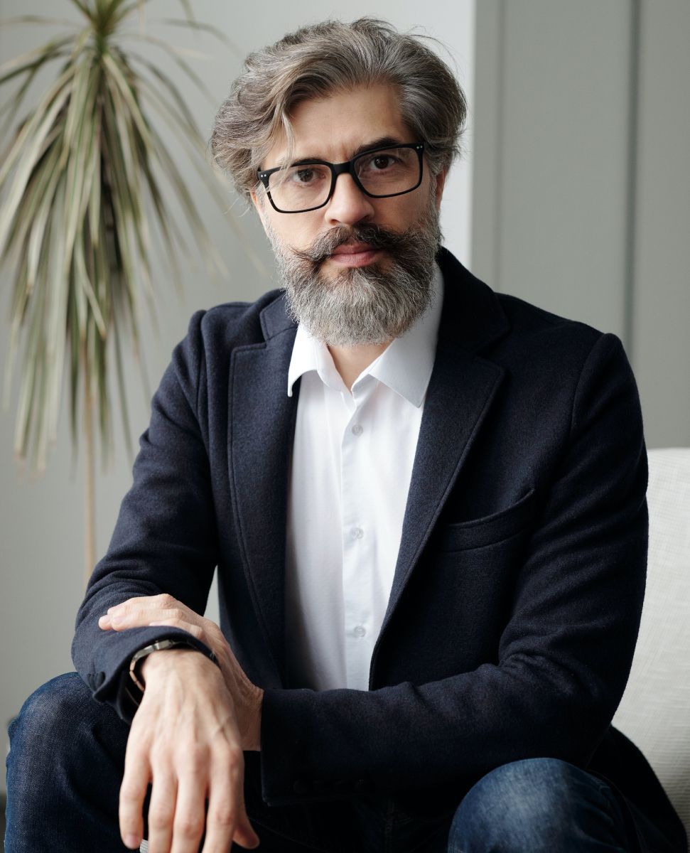 Man with beard, glasses, in a blazer and button-down shirt, seated indoors, looking at the viewer.