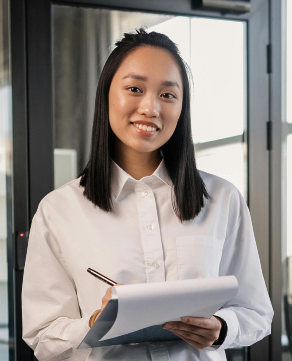 Woman in white shirt smiles, holding clipboard and pen, near a glass door.