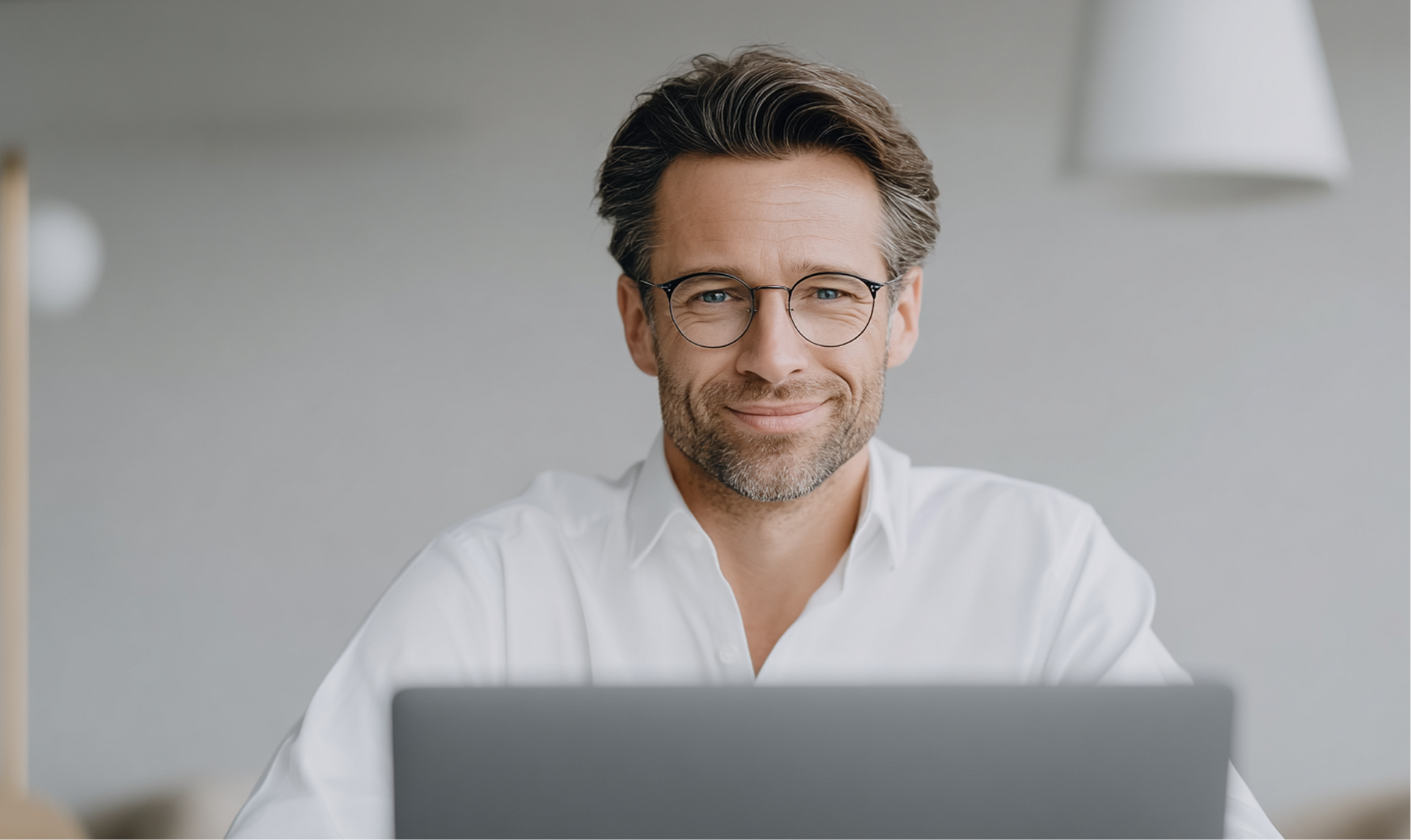 Un uomo con gli occhiali sorride alla telecamera mentre lavora al computer portatile. Sfondo chiaro.