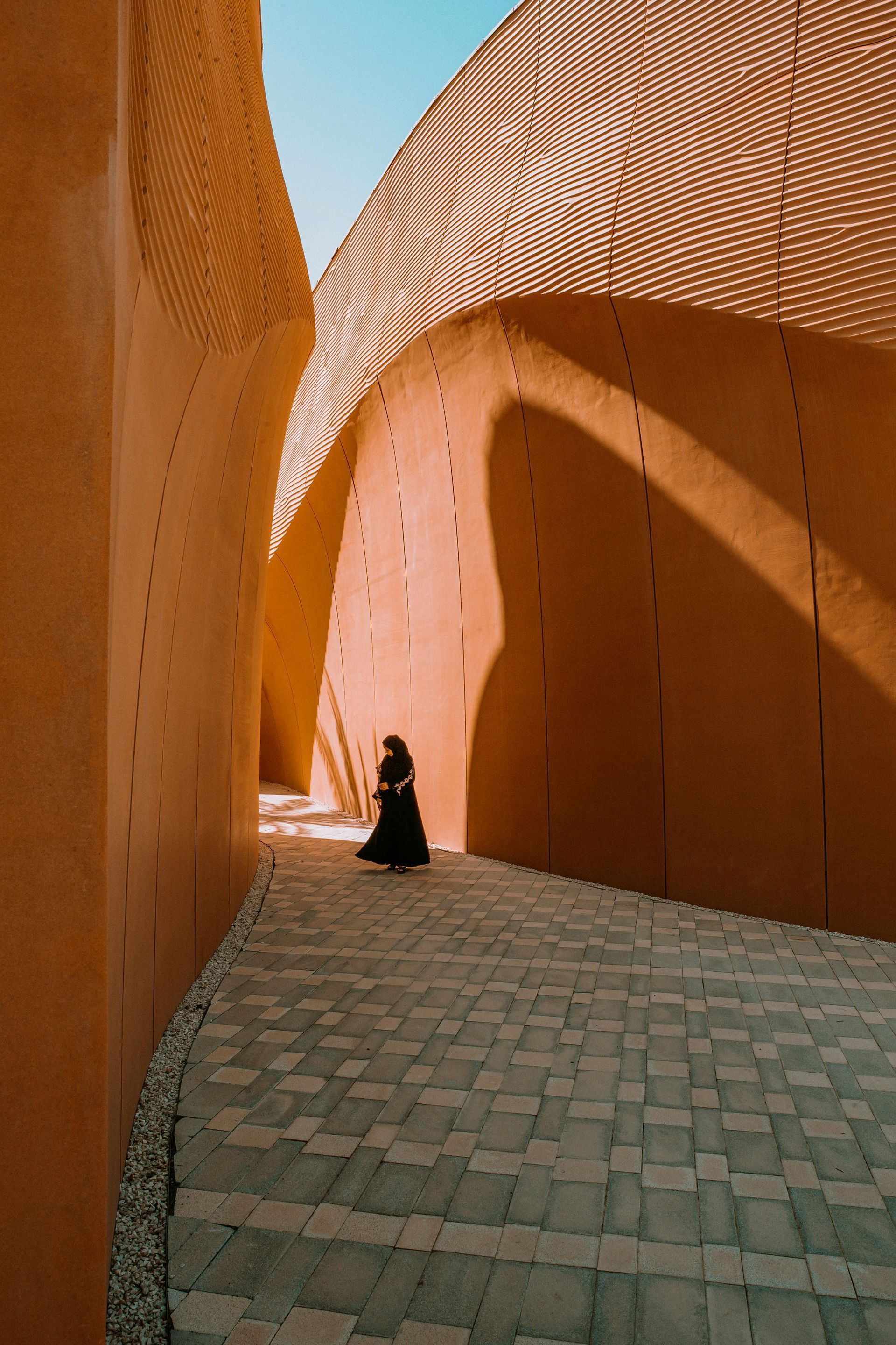 A person walks along a narrow, textured, sunlit pathway between tall, tan walls.
