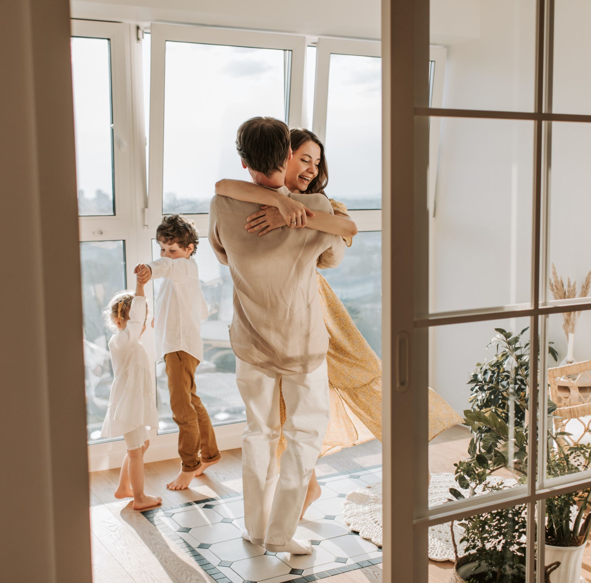 Family embracing and dancing in a sunlit room with large windows; children dancing nearby.