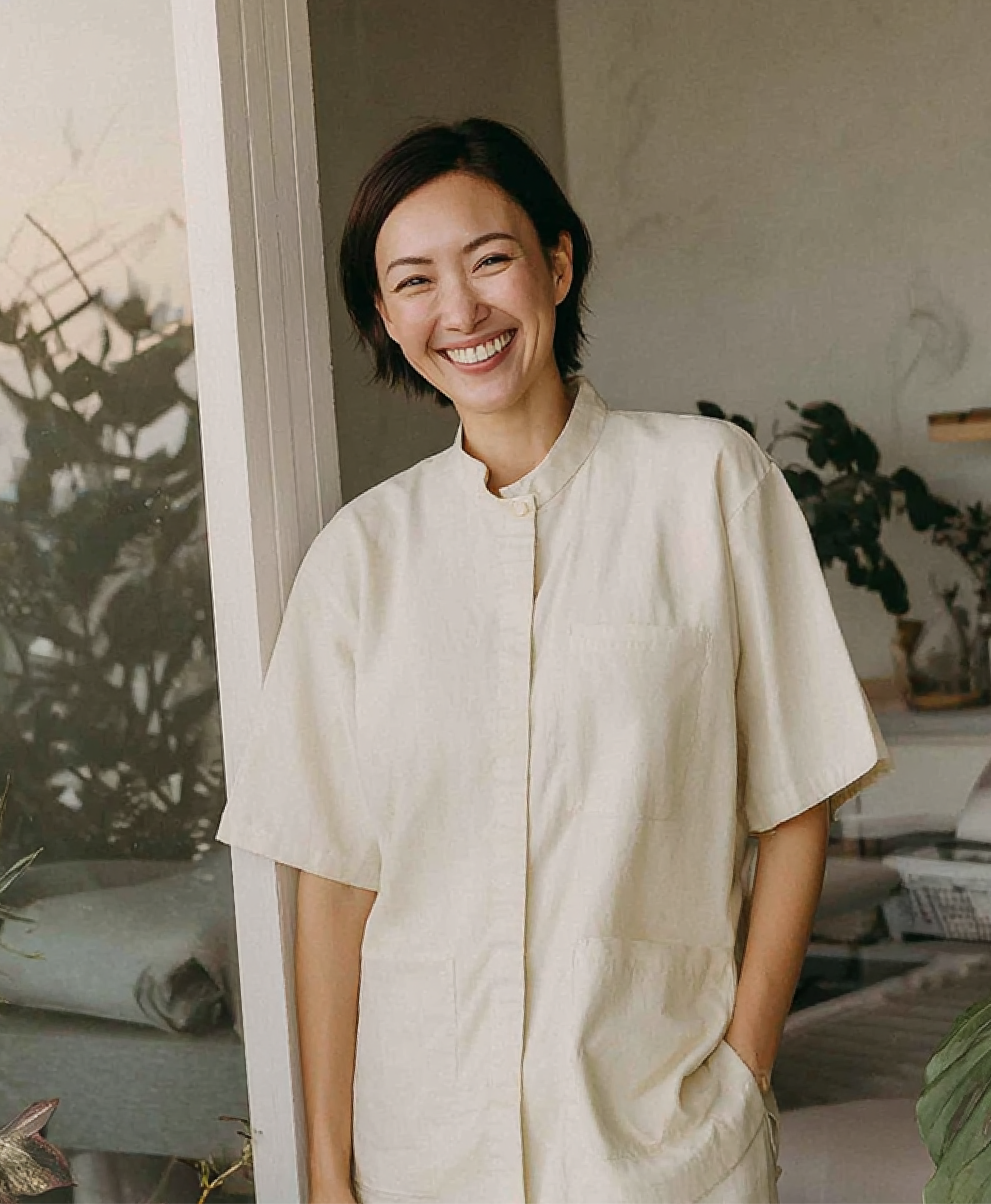 Woman smiles, wearing a light-colored short-sleeved jumpsuit, standing indoors near a window with sunlight.