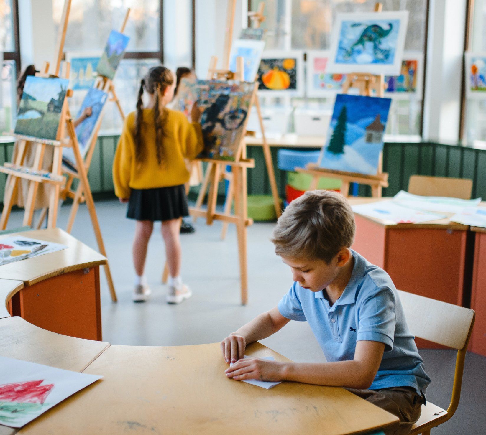 A boy is sitting at a table in a classroom while a girl stands in the background.