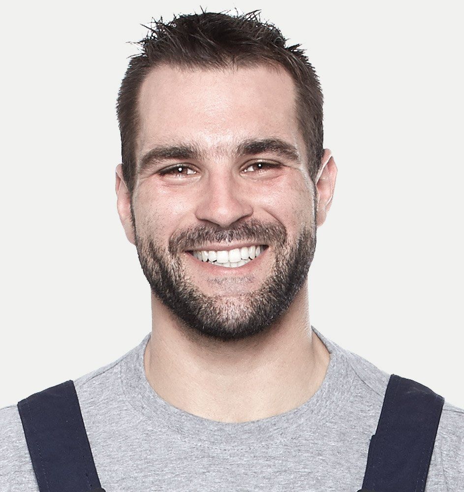 Man with dark hair and beard, smiling. Wearing a grey shirt with blue straps, against a white backdrop.