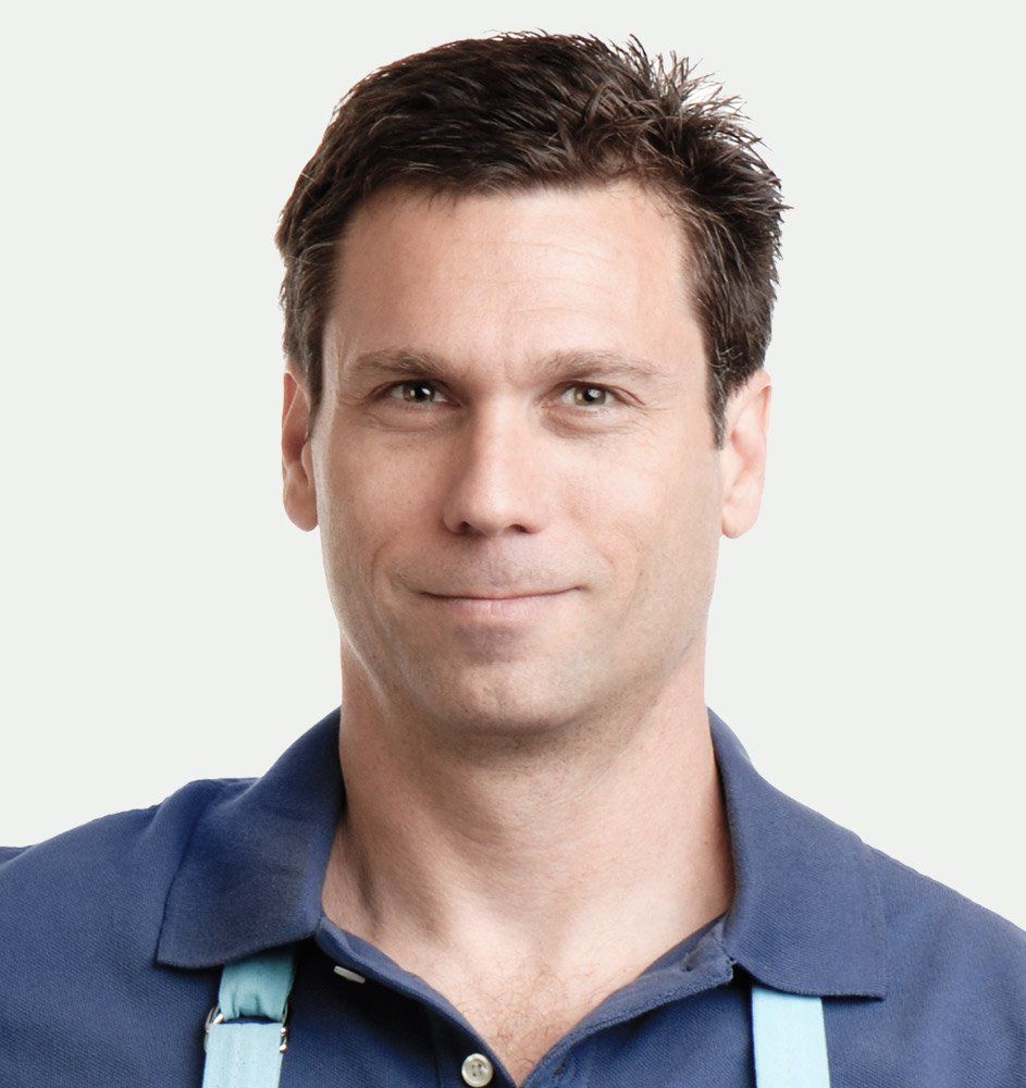 Man with dark hair and blue shirt smiles, wearing a light blue apron against a white background.