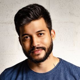 Portrait of a smiling man with dark hair and beard in a blue shirt against a light wall