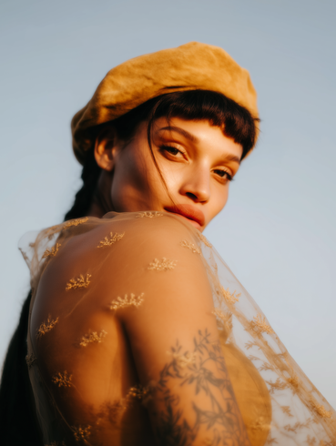 Portrait of a person in a mustard beret and sheer gold top against a soft blue background