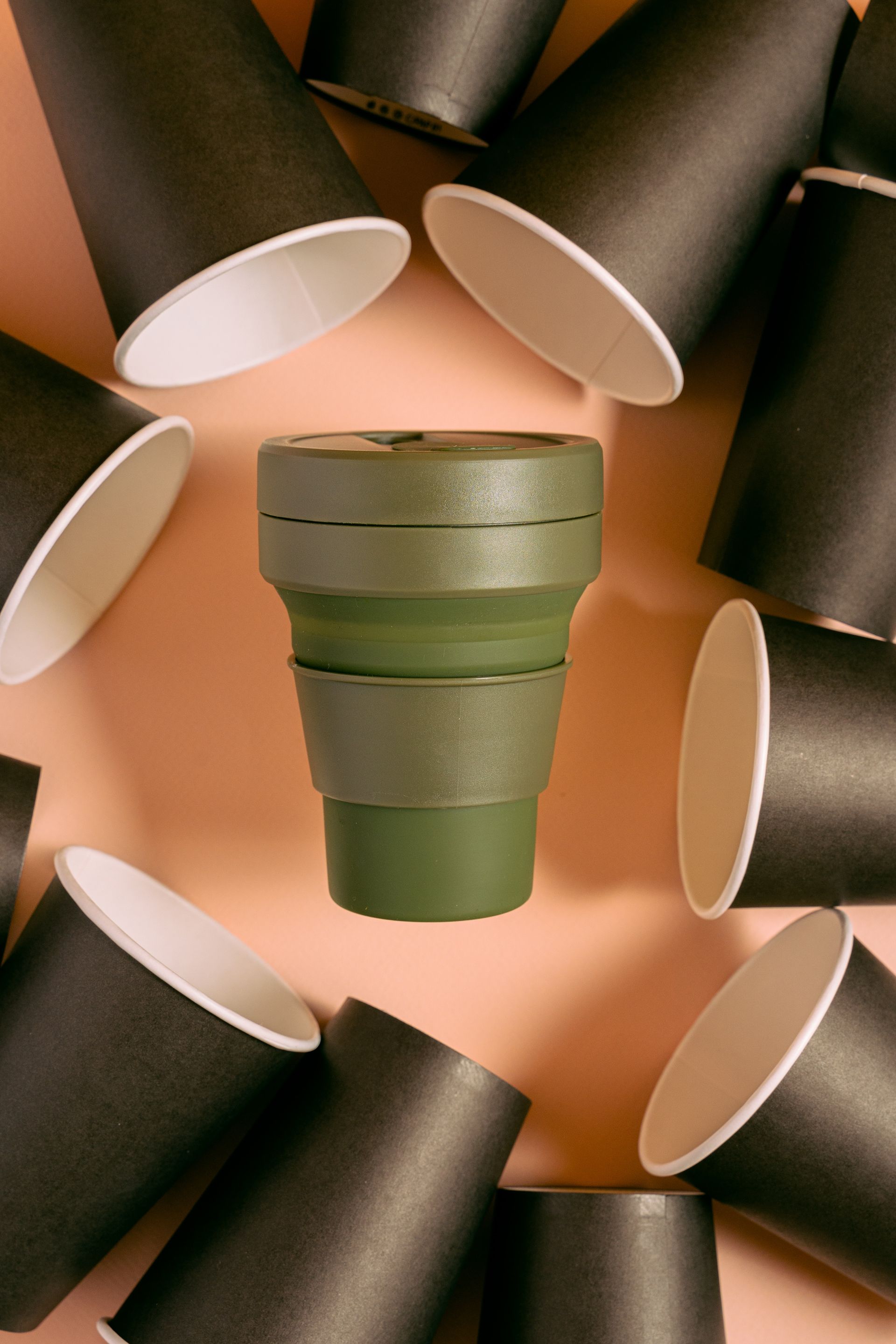 Green military-style mug centered among black paper cups on a warm peach background